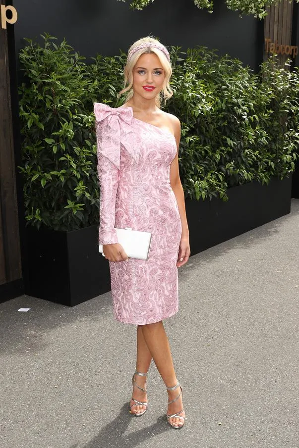 Woman in pink one-shoulder floral dress, headband, holding a white clutch, outdoors.