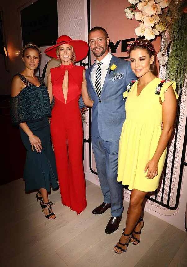 Four people posing together; woman in red outfit and hat, man in blue suit, women in green and yellow dresses.