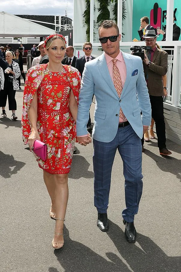 A couple walks in formal attire; woman in floral red dress, man in blue suit, holding hands at an outdoor event.