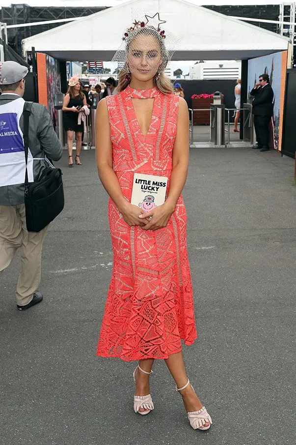 Woman in a red patterned dress with headpiece, holding a "Little Miss Lucky" book at an outdoor event.