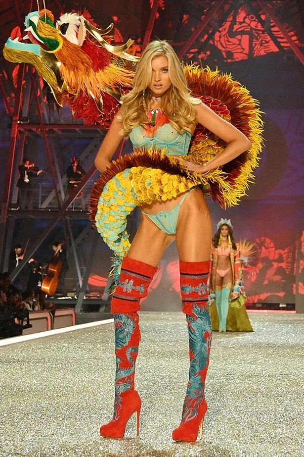 Model on runway in colorful dragon-themed outfit with high red boots, sparkly background.