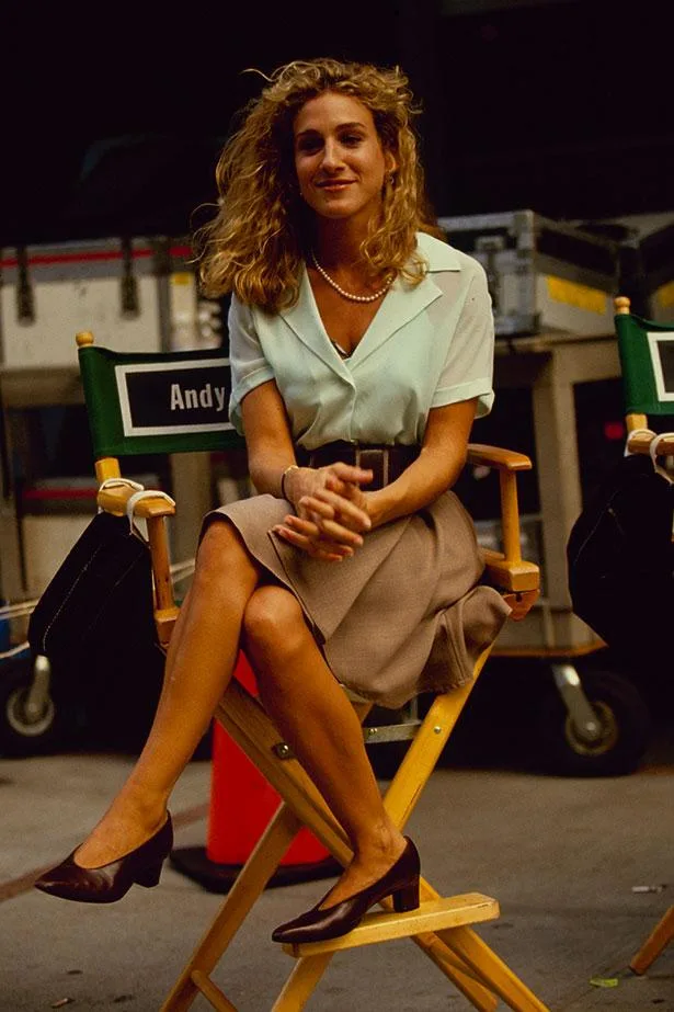 Young woman with curly hair sitting on a director's chair labeled "Andy," wearing a blouse, skirt, and heels.