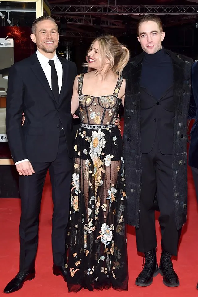 Three people smiling on a red carpet, one woman in a floral dress between two men in suits.