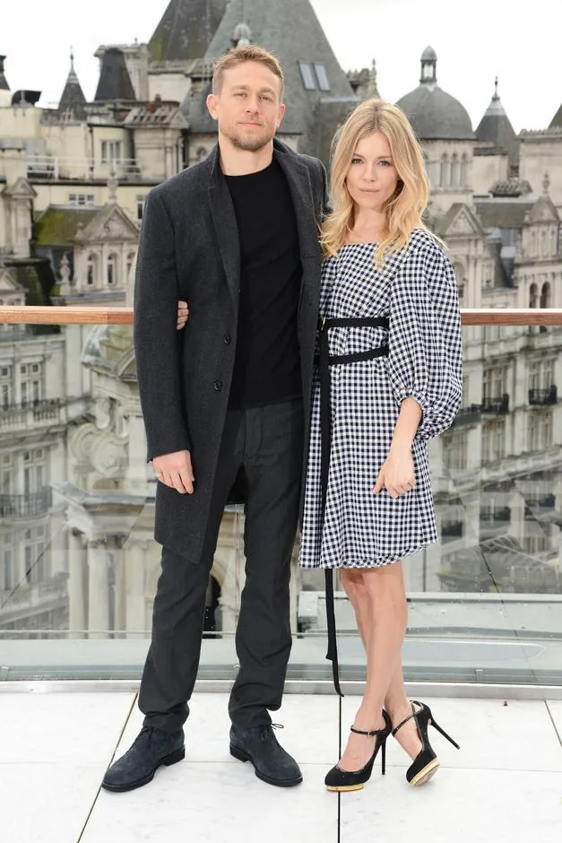 Man in dark coat and woman in checkered dress posing on rooftop with cityscape background.