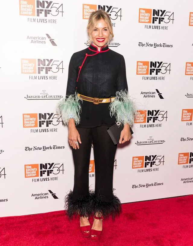 A person in a black outfit with feather details poses on a red carpet at NYFF54, with event logos in the background.