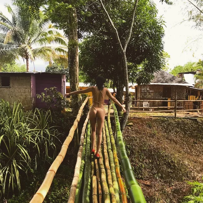A person in a swimsuit walks on a bamboo bridge with tropical greenery and huts in the background.