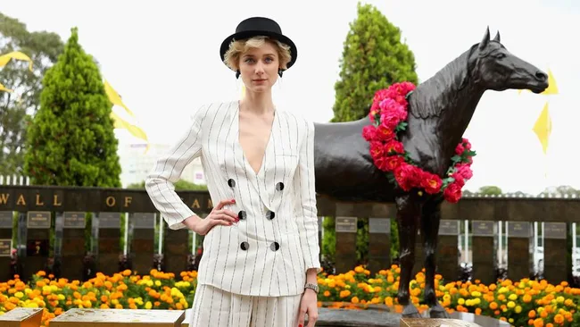Person in striped suit and black hat stands by horse statue adorned with flowers.