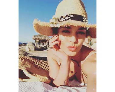 Vanessa Hudgens in a straw hat with floral detail, lying on a beach towel under the sun, pouting at the camera.