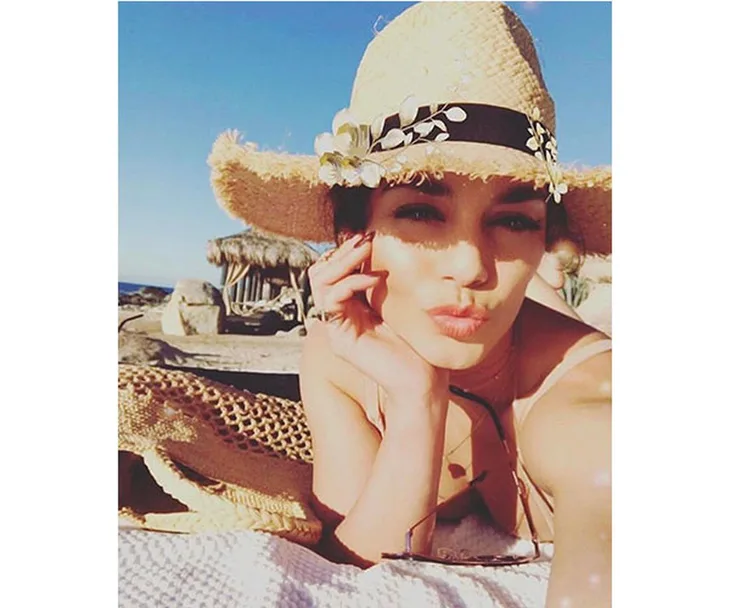 Vanessa Hudgens in a straw hat with floral detail, lying on a beach towel under the sun, pouting at the camera.