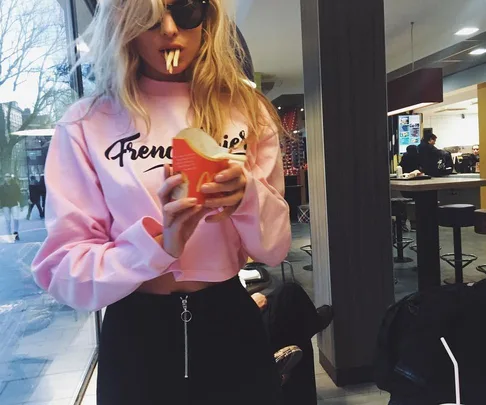 Young woman in pink sweater playfully holding fries in her mouth while standing in a fast-food restaurant.