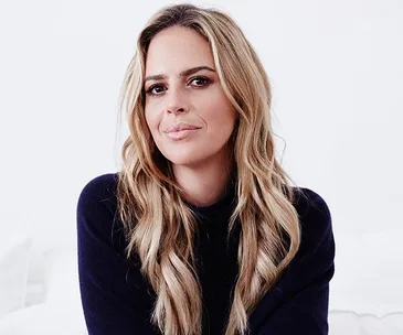 "A woman with long blonde hair, wearing a dark sweater, smiles softly while looking into the camera against a white background."