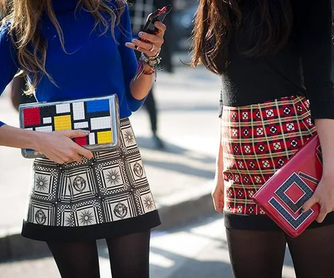 Two stylish women are standing, each holding patterned clutches, one in blue top and geometric skirt, the other in black top and red skirt.