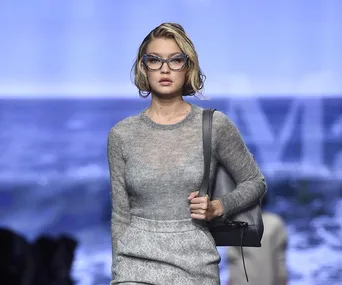 Model in gray sheer top and skirt, blue glasses, carrying a handbag, walking on a runway with a blurred ocean background.