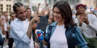 Woman in blue jacket holds a Pepsi can, facing a crowd of cheering people.