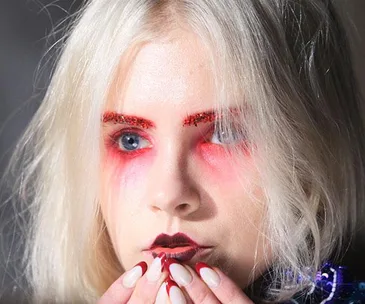 Model with bold red eye makeup, glitter eyebrows, dark lips, pale blonde hair, and manicured nails at MBFWA 2017.