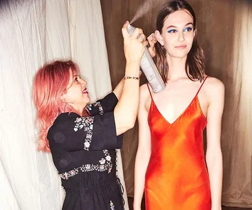 Hairstylist sprays model's hair backstage; model in orange dress with bright blue eye makeup.