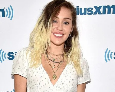 Miley Cyrus at SiriusXM event with shoulder-length ombre hair, wearing a white floral dress and layered necklaces.