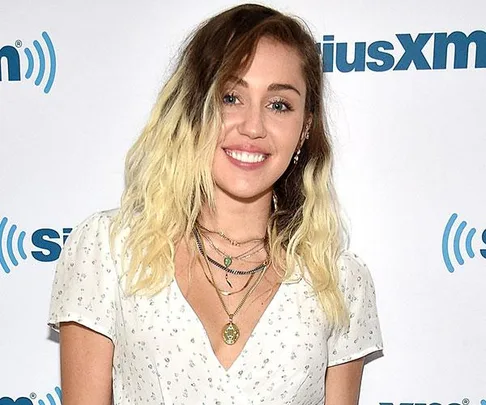 Miley Cyrus at SiriusXM event with shoulder-length ombre hair, wearing a white floral dress and layered necklaces.