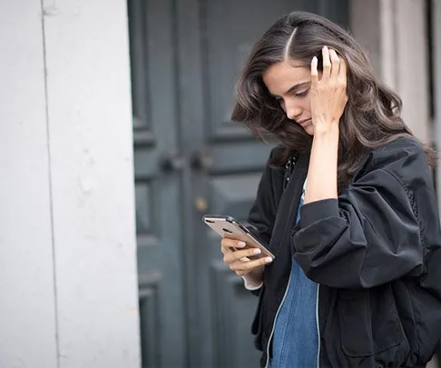 A woman looks at her smartphone while standing outside, touching her hair with a concerned expression.