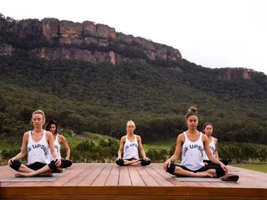 Group of women wearing "The Upside" tank tops practice yoga outdoors on a wooden platform with a mountainous forest backdrop.