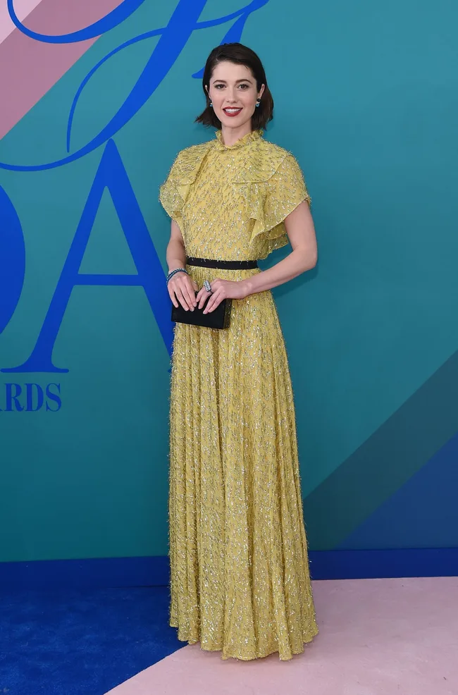 Person in a sparkling yellow dress holding a black clutch, standing against a blue and green background.
