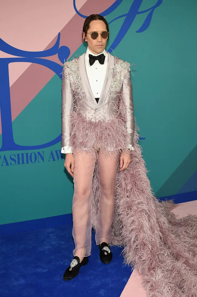 Person in a pink feathered suit with a long train, wearing sunglasses, stands on a blue carpeted event backdrop.