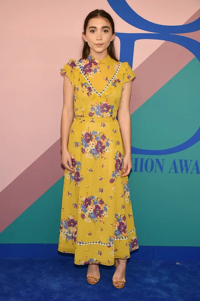 Young woman in a yellow floral dress stands on a blue carpet at a fashion awards event.