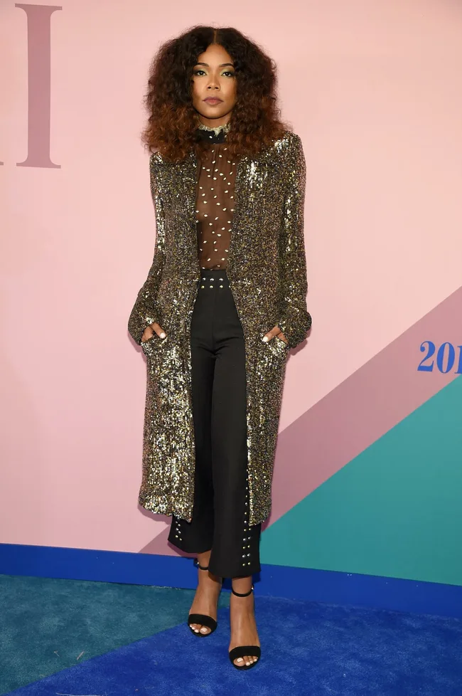 Person in sequined coat and sheer polka dot top on blue carpet, pink and teal background.