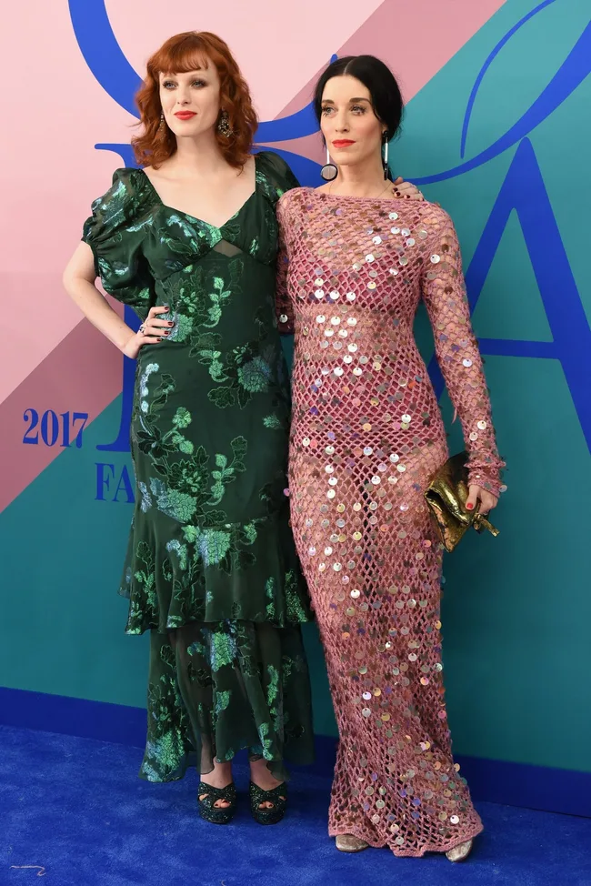 Two women pose at a fashion event; one in a green floral dress, the other in a pink sequin net dress holding a clutch.