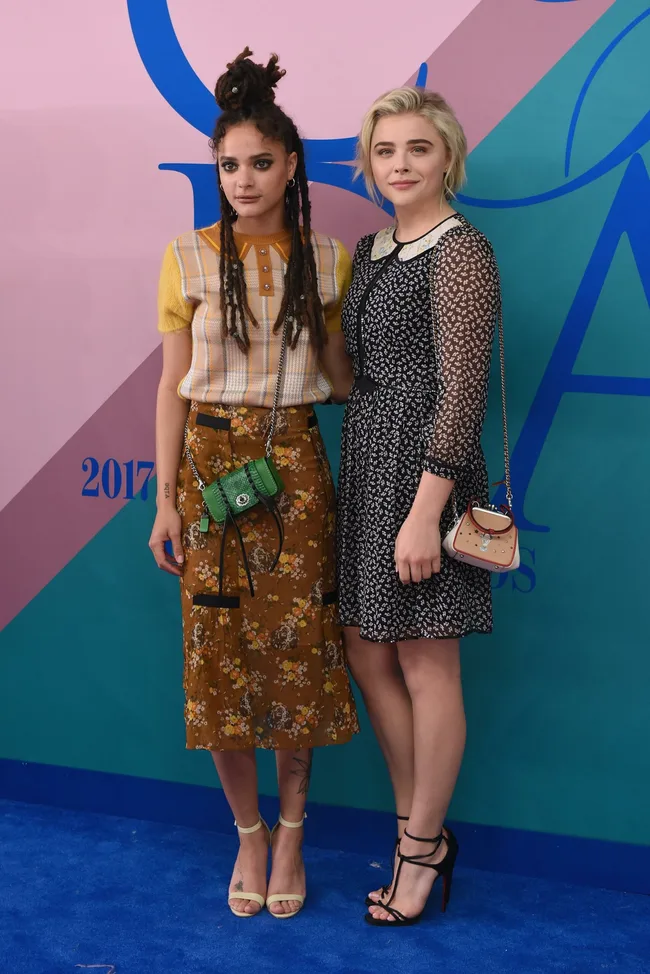 Two women posing together at a fashion event, wearing patterned outfits and carrying small handbags.