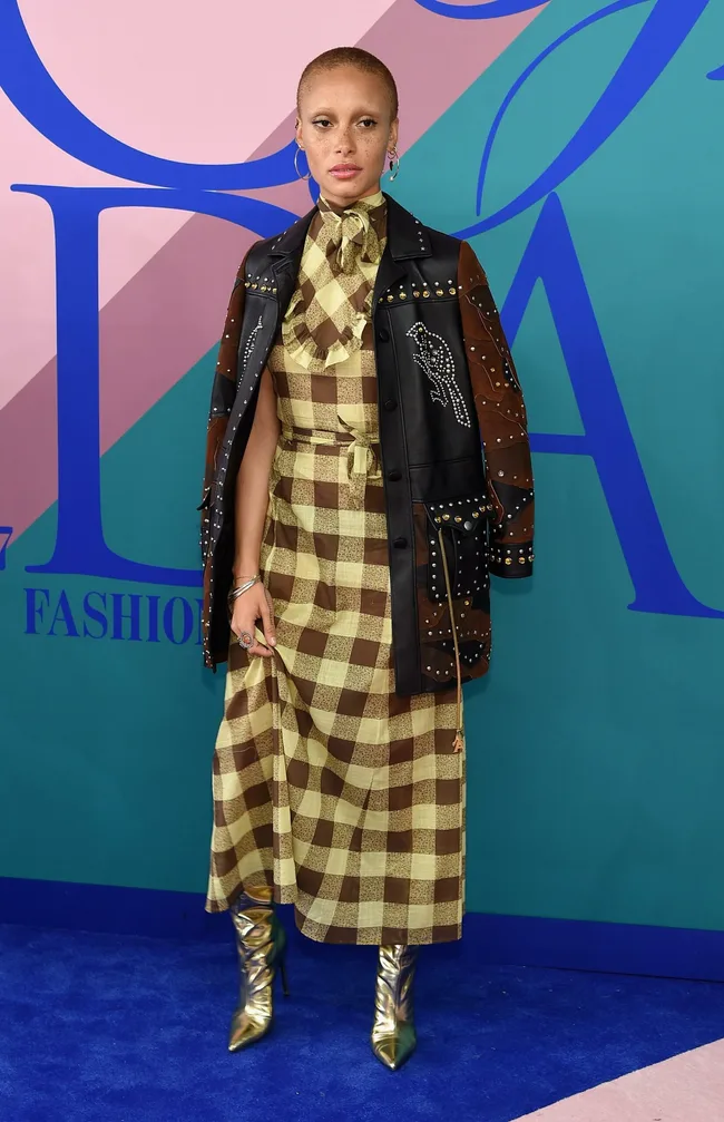 Person in checkered dress and studded jacket on the blue carpet at a fashion event.