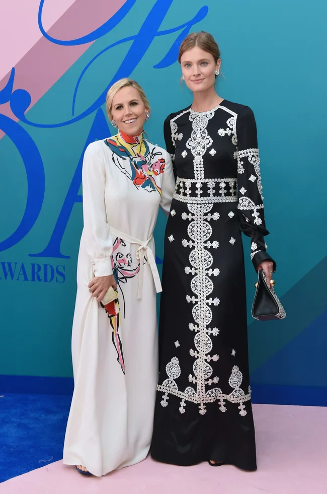 Two women in elegant dresses pose together on a colorful, patterned backdrop at an event.