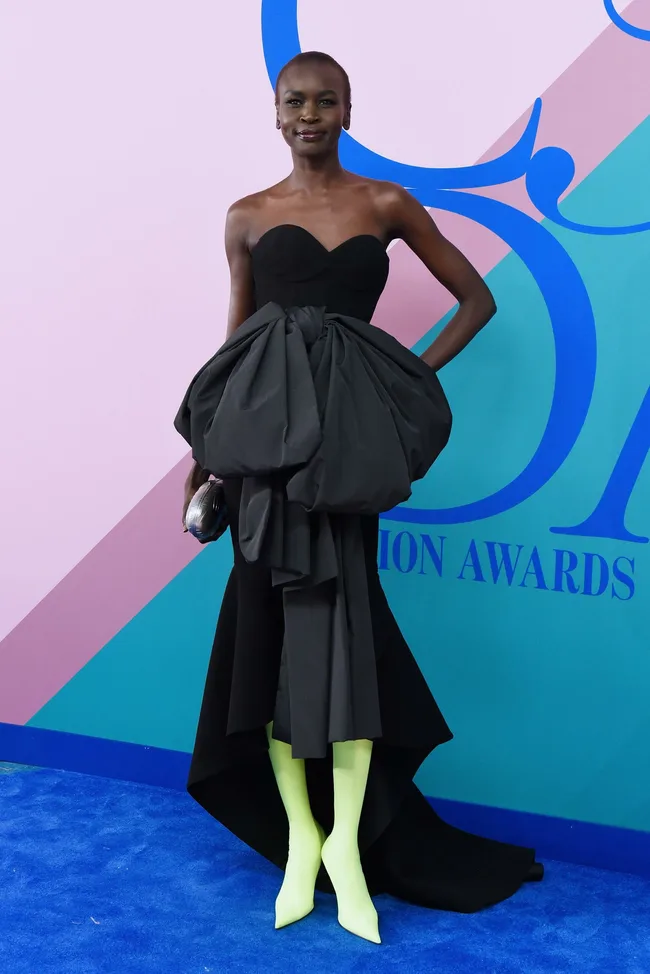 Model in a strapless black dress and neon yellow boots poses on blue carpet at a fashion event.