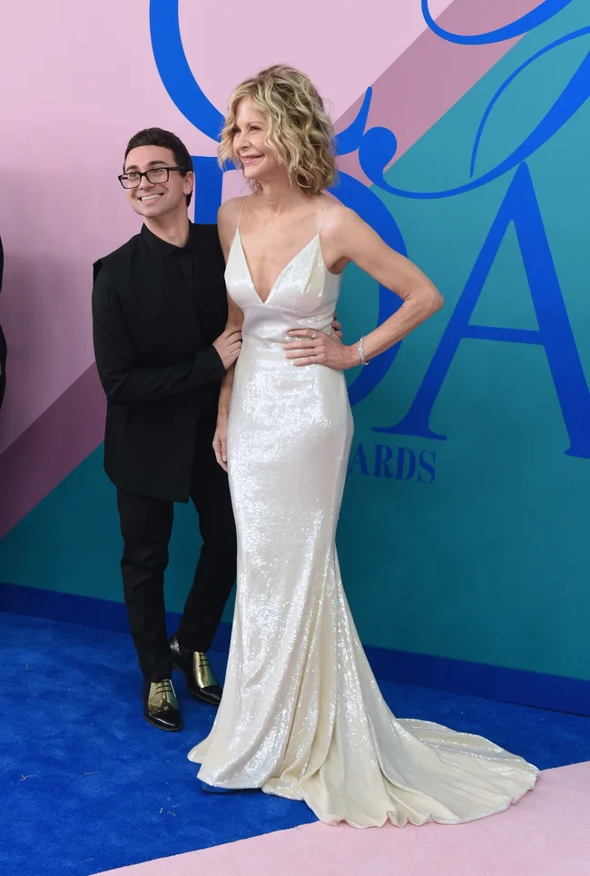 Man and woman in evening wear on a red carpet; woman in white gown, man in black suit with gold shoes.