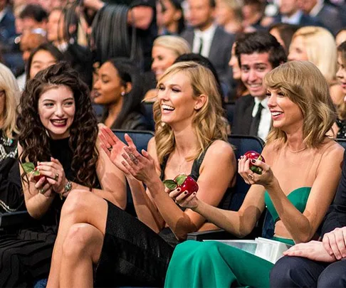 Three women, including Lorde, holding apples and laughing in a seated crowd at an event, with others around them also smiling.