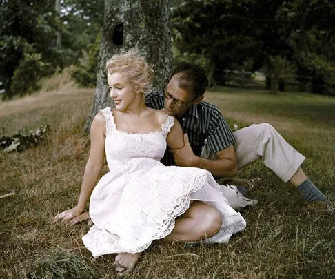 Marilyn Monroe sitting in a park wearing a white dress, leaning against a tree with a man wearing glasses beside her.