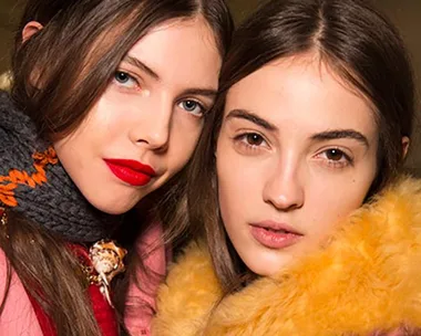 Two women in winter coats and scarfs, one with red lips while other natural look, representing winter fashion trends.