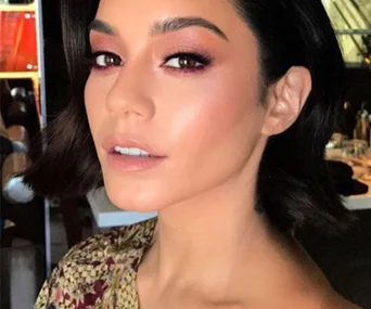 A person with short dark hair and matching eyeshadow and lipstick, wearing a floral top, looking towards the camera.