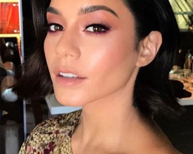 A person with short dark hair and matching eyeshadow and lipstick, wearing a floral top, looking towards the camera.