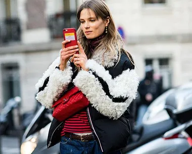 Woman in a black and white fur coat using a red smartphone on a city street.