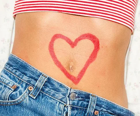 Close-up of a woman's midriff with a heart shape drawn in red on the skin, wearing a striped top and jeans.