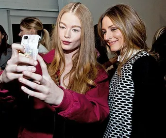 Two women taking a selfie in a social setting, one with long red hair in a red jacket, the other with long brown hair smiling.