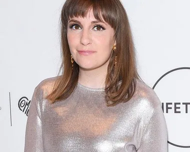 Lena Dunham with shoulder-length brown hair and bangs, wearing a shiny silver top, standing in front of a white backdrop.