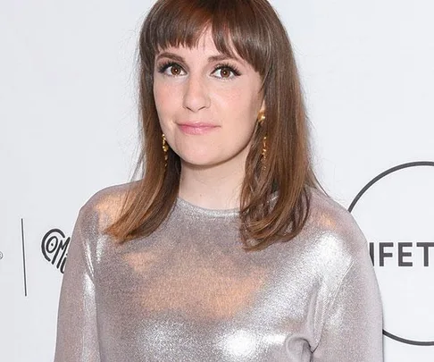 Lena Dunham with shoulder-length brown hair and bangs, wearing a shiny silver top, standing in front of a white backdrop.