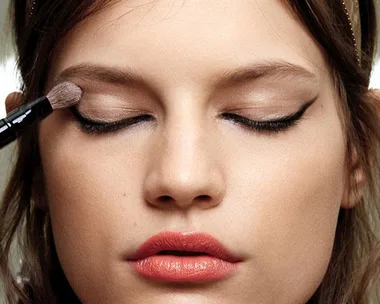 A close-up of a woman with closed eyes, applying eyeshadow with a brush. She has winged eyeliner and glossy lips.