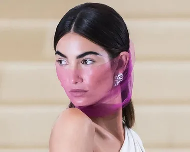 Lily Aldridge with a pink sheer scarf partially covering her face, wearing a white one-shoulder dress and diamond earrings.