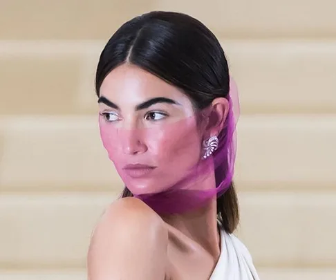 Lily Aldridge with a pink sheer scarf partially covering her face, wearing a white one-shoulder dress and diamond earrings.