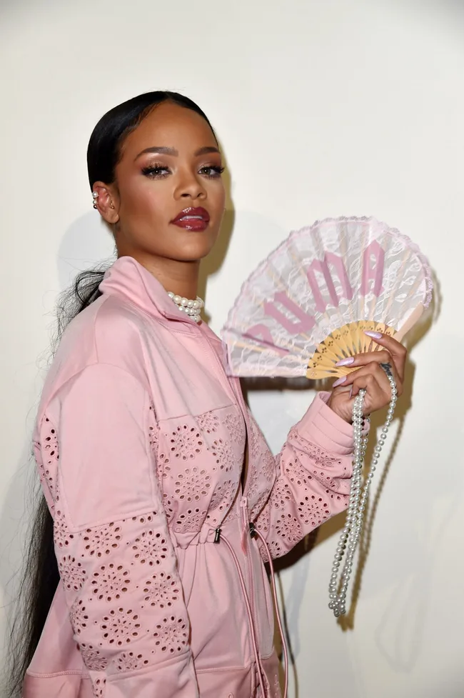 Young woman in pink jacket holding a lace fan with "PUMA" text, wearing pearl jewelry, looking stylishly at the camera.