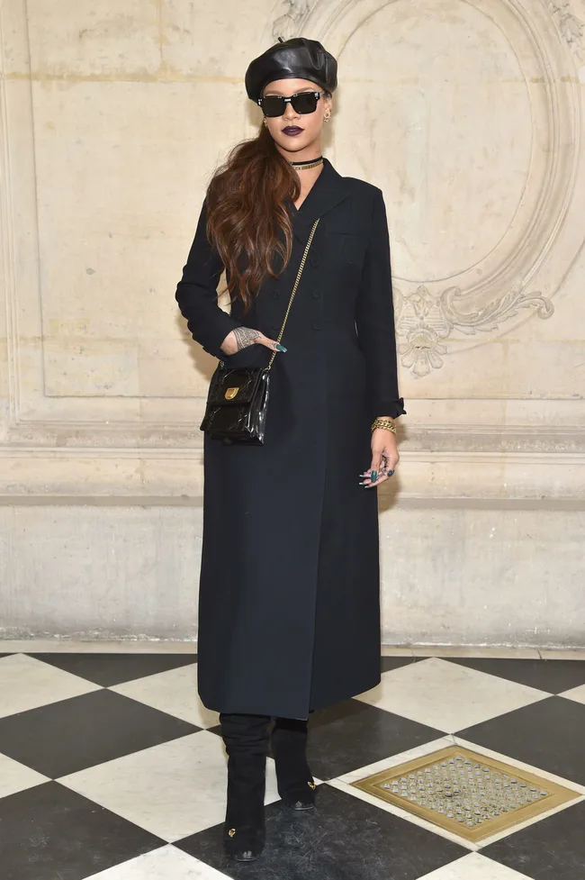 Person in black coat, beret, and sunglasses, standing on checkered floor with a black handbag.