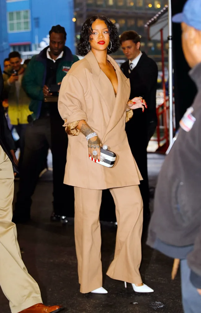 Fashionably dressed individual in a tan outfit with red lipstick and jewelry, holding a small bag, on a city street.
